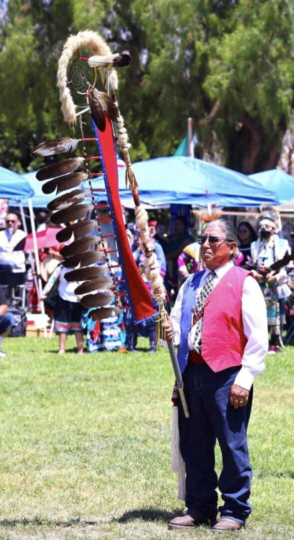 Man in traditional attire holding a feather staff at a cultural gathering.