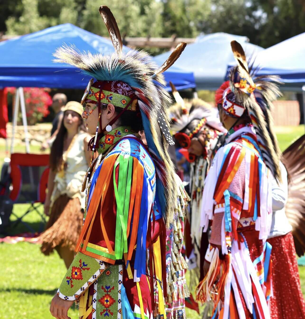 Two Native American dancers in colorful traditional regalia at an outdoor event.