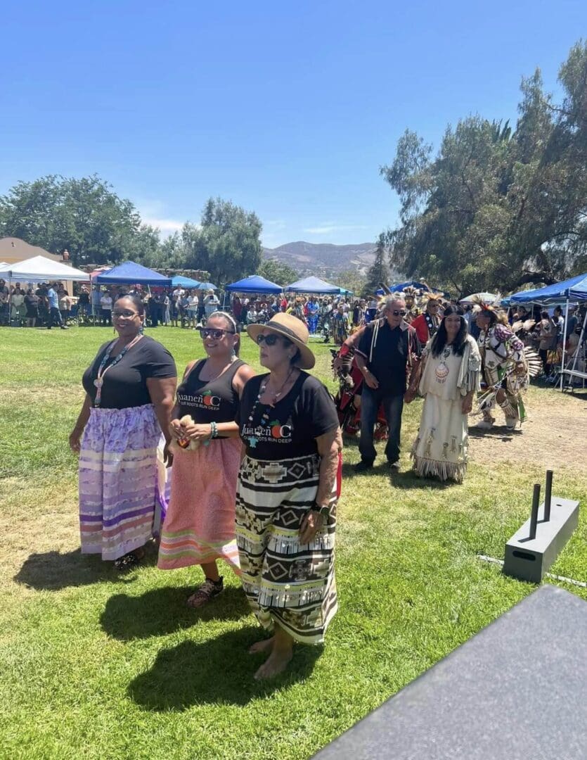 People gather outdoors at a sunny market or festival with tents and traditional clothing.