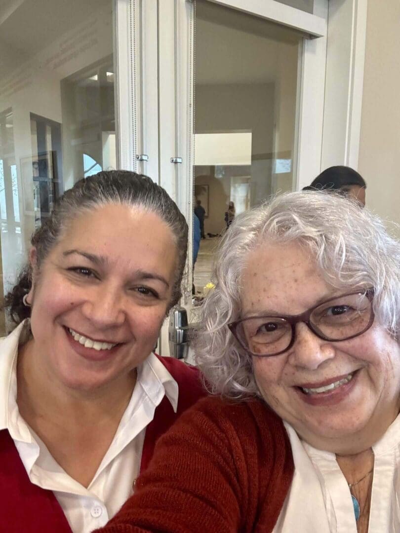 Two women smiling indoors, wearing glasses.